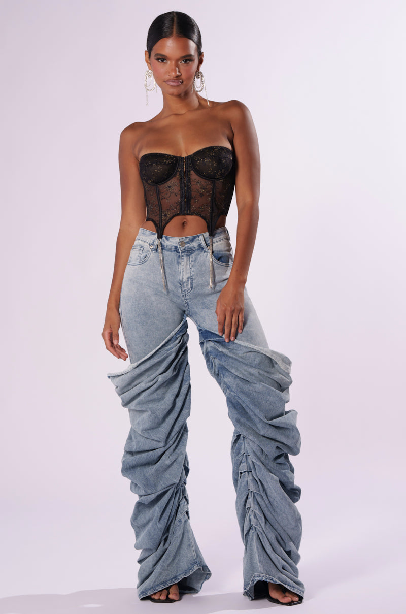 A woman stands against a plain background, wearing a black lace strapless top and OH YEAH BABY DENIM PANT by JCR Trading LTD with an exaggeratedly ruched baggy effect. She has dark hair pulled back, large hoop earrings, and an assertive pose with one hand on her hip and the other relaxed by her side.