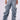 Someone is wearing the OH YEAH BABY DENIM PANT by JCR Trading LTD, featuring highly ruched, baggy light blue denim with a mid-rise waist. They stand against a plain white background with their hands resting at their sides. The jeans showcase exaggerated, dramatic folds and are paired casually with sandals.