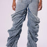 Someone is wearing the OH YEAH BABY DENIM PANT by JCR Trading LTD, featuring highly ruched, baggy light blue denim with a mid-rise waist. They stand against a plain white background with their hands resting at their sides. The jeans showcase exaggerated, dramatic folds and are paired casually with sandals.