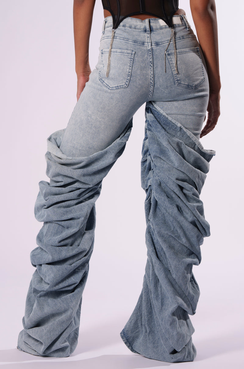 Person wearing OH YEAH BABY DENIM PANT from JCR Trading LTD in a light-washed, extremely oversized style paired with a black top. The mid-rise waist jeans are gathered at the ankles, creating a billowing, ruched baggy effect. The back pockets and belt loops are visible against the white background.