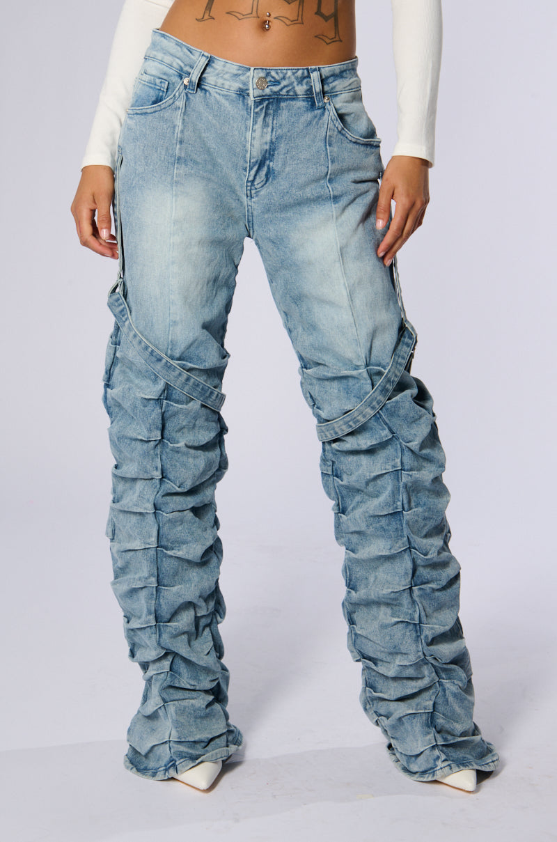 A person is shown from the torso down, wearing the GOING CRAZY DENIM PANT WITH BUCKLE—a pair of light blue, high-waisted, ruched denim jeans paired with white pointed-toe high heels. The jeans feature a unique design with gathered pleated details and an adjustable five-pocket layout. The person is posing with hands in pockets.