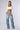 A woman stands confidently against a plain background, wearing a white crop top that reveals a tattoo on her stomach reading "1999". She pairs it with the GOING CRAZY DENIM PANT WITH BUCKLE, featuring light-wash, bulky, ruched denim and adjustable denim straps in addition to a five-pocket design. She completes the look with white pointed-toe heels, a chunky necklace, and earrings.