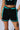 A person wearing black BALLIN ALL NIGHT SHORT with turquoise trim and a matching top stands confidently. A striking tattoo of the number 1999 adorns their stomach, complemented by a skull tattoo on their thigh. Their hand rests on the shorts, emphasizing the adjustable drawstrings, while parts of their upper body and legs are clearly visible.