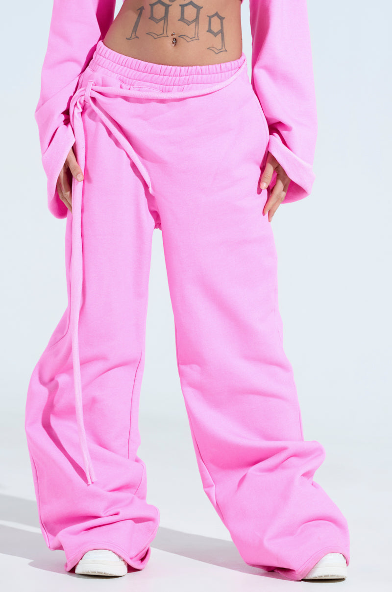 Wearing an athleisure ensemble, the person showcases oversized pink ISSA WIDE LEG JOGGER sweatpants with a striking long tie detail, paired with a matching cropped top. A "1999" tattoo adorns their stomach while white sneakers complete the look.