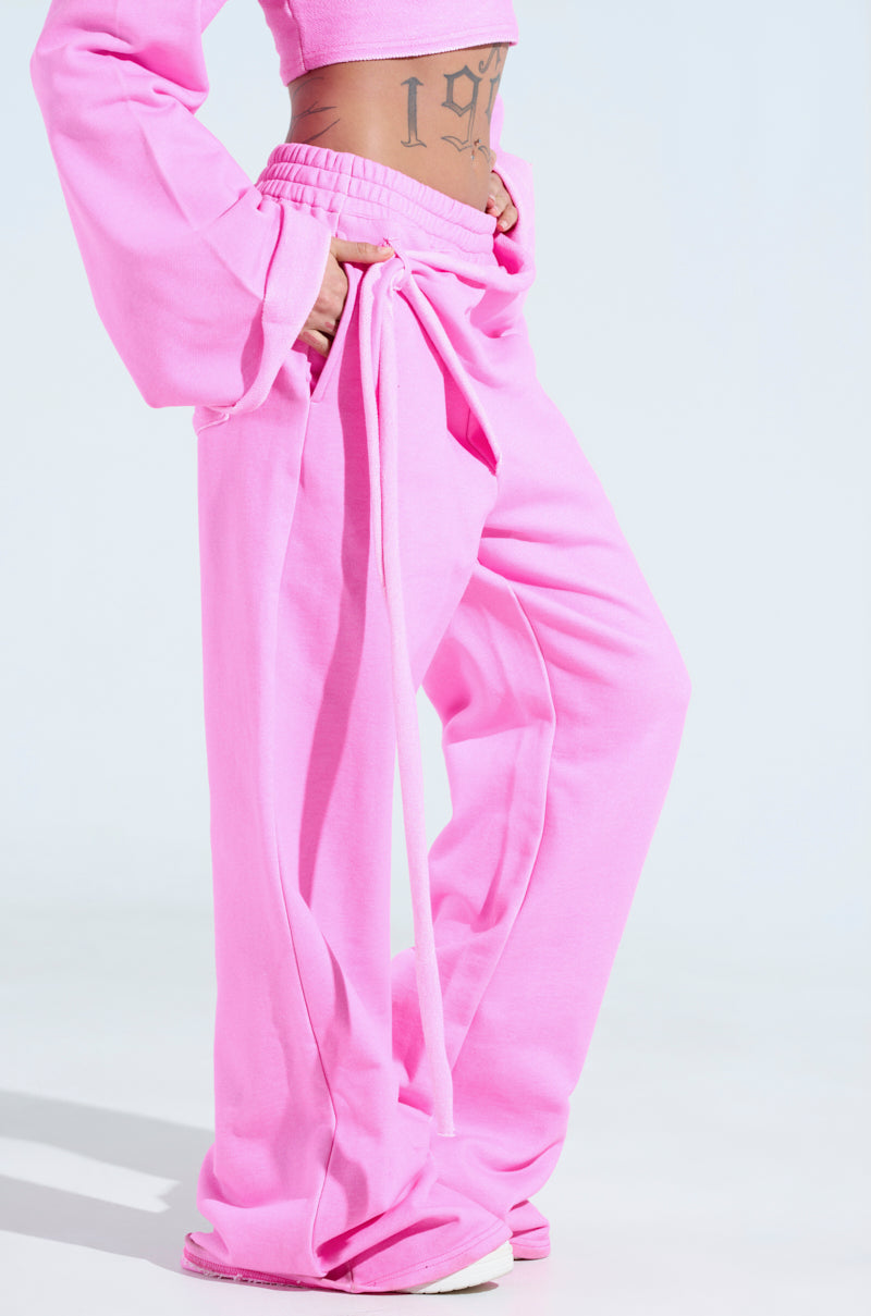 The photo showcases someone in athleisure, wearing ISSA WIDE LEG JOGGER and a long-sleeved, pink cropped top. Their tattooed abdomen peeks above white sneakers. The light blue background enhances the vibrancy.