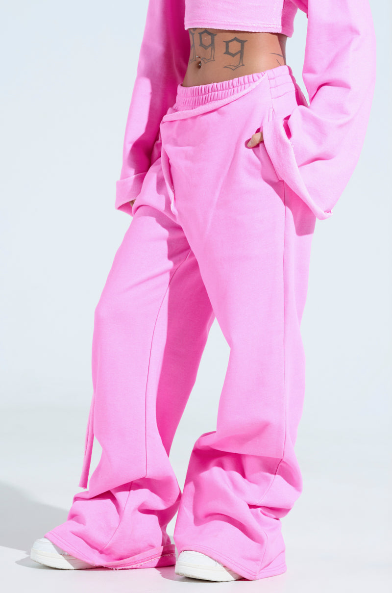 Wearing the bright pink Issa Wide Leg Jogger with a matching long-sleeve crop top, showcasing a midriff tattoo, the cotton joggers' elastic waistband pairs perfectly with white sneakers, making the athleisure outfit pop against the plain light-colored background.