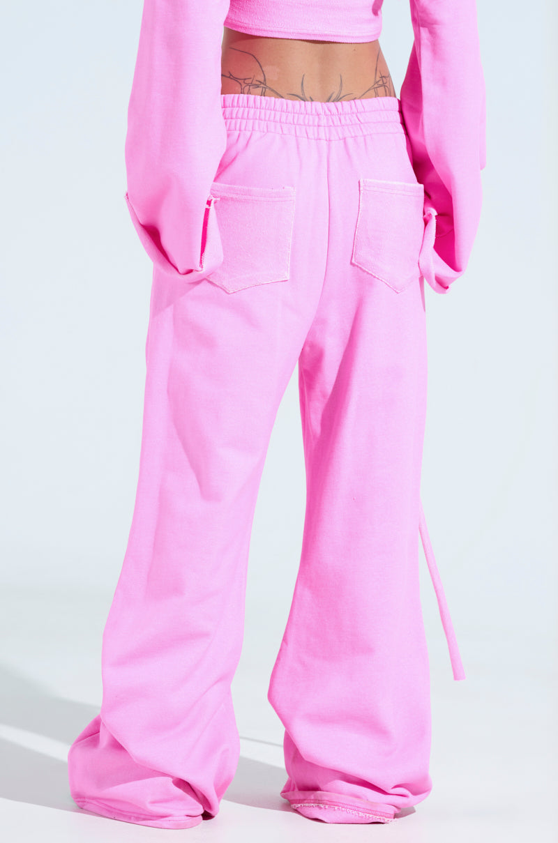 The person is wearing an athleisure ensemble featuring loose, bright pink ISSA WIDE LEG JOGGER Pants with patch pockets on the back, paired with a matching long-sleeve top. The outfit pops against the plain light background.