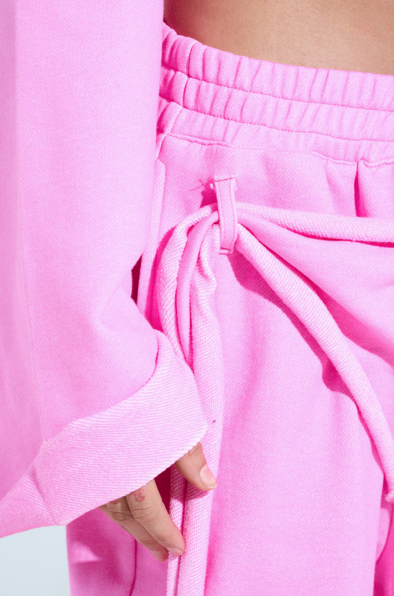 A close-up shows someone in a bright pink cotton ensemble, featuring the ISSA WIDE LEG JOGGER with an elastic waistband and side ties. Their hand softly rests near the pocket, accentuating the athleisure's soft texture.