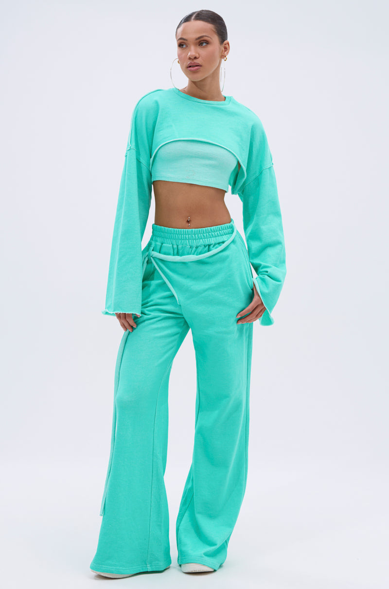 A woman poses against a plain background wearing the ISSA JOGGER PANT IN TIFFANY, styled with wide-leg sweatpants and a matching long-sleeve crop top layered over a bandeau. She completes the look with large hoop earrings. 0