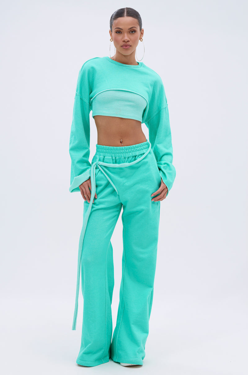 A woman stands confidently in the ISSA JOGGER PANT IN TIFFANY, featuring wide-leg joggers with a high-rise elastic waistband and a matching aqua blue long-sleeve cropped top. She accessorizes with hoop earrings and has her hair pulled back. 6