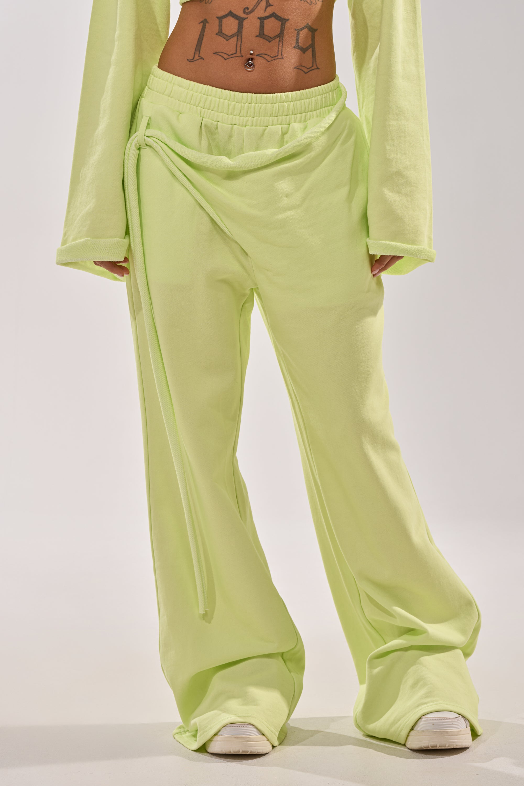 Wearing the TRY ME BABY WIDE LEG JOGGER in lime with a high-rise elastic waistband and wrap detail, paired with a matching long-sleeve top. White shoes complete the look, revealing a “1999” midriff tattoo against a plain light background. 1