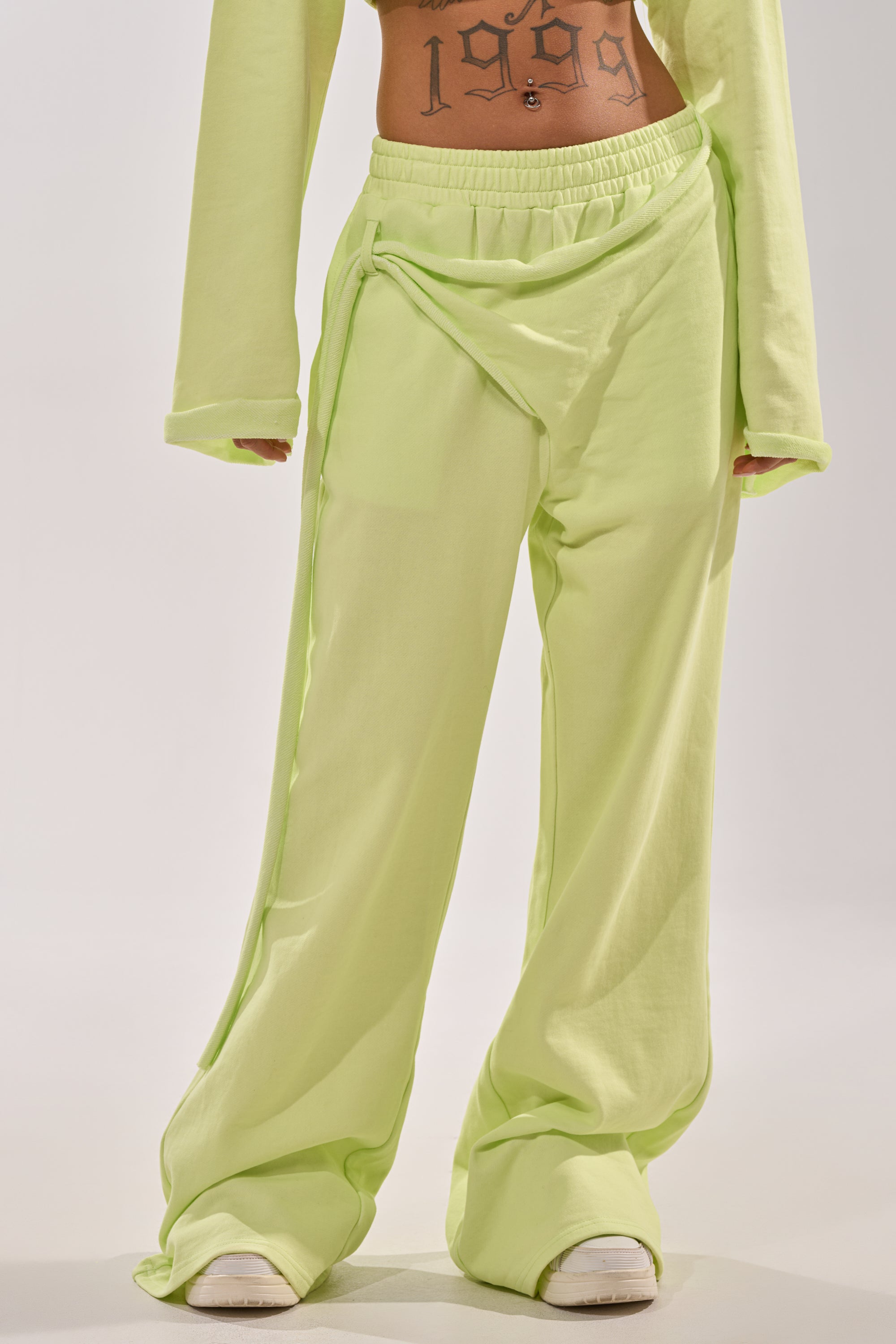 Person wears the TRY ME BABY WIDE LEG JOGGER in light green with a matching long-sleeve top, revealing a "1999" midriff tattoo. They stand against a plain background, white shoes partially visible. 2
