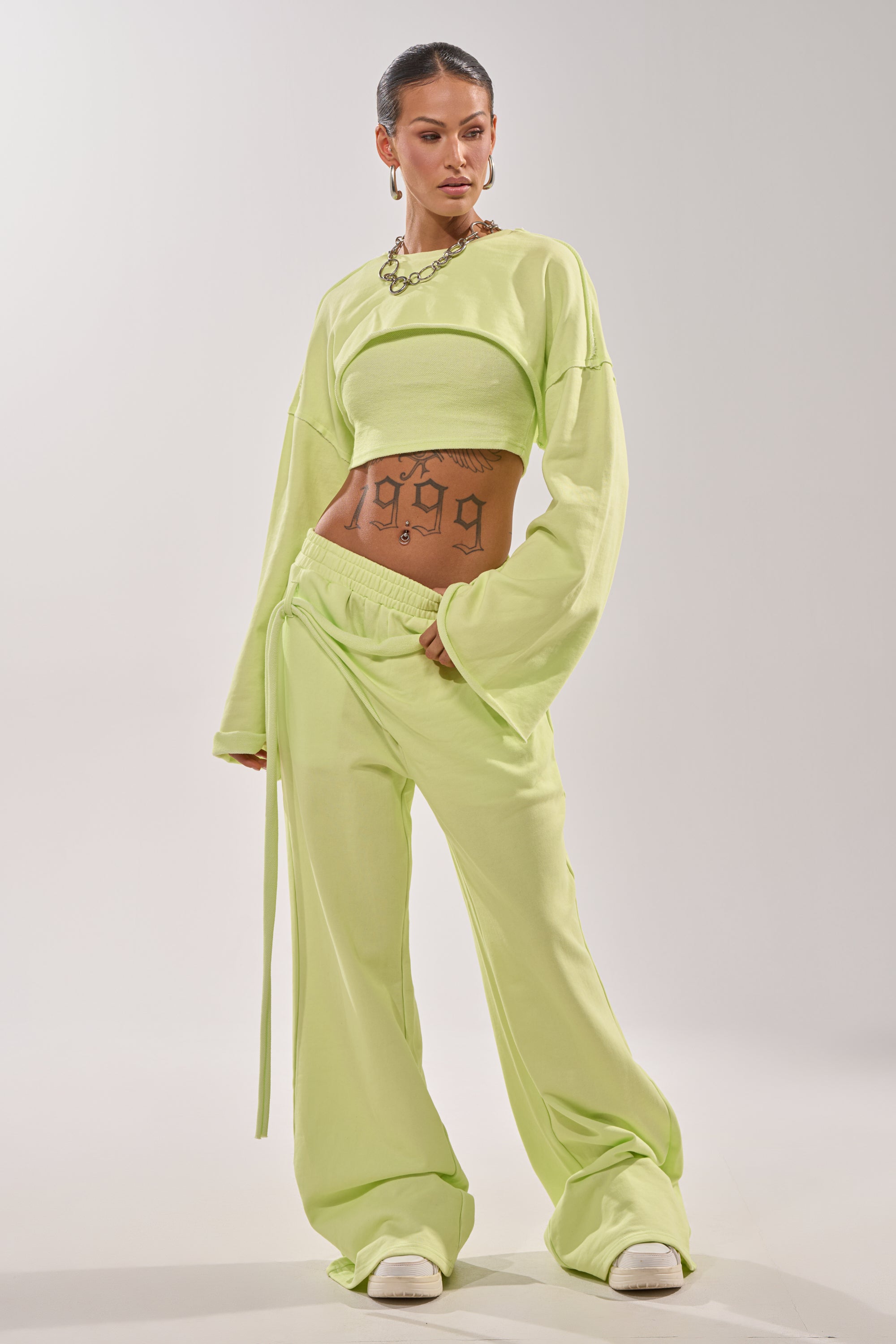 A woman strikes a confident pose in the TRY ME BABY WIDE LEG JOGGER, paired with a light green crop top and chunky chain necklace. Her bold "1989" tattoo and white sneakers complete the effortless streetwear look. 6