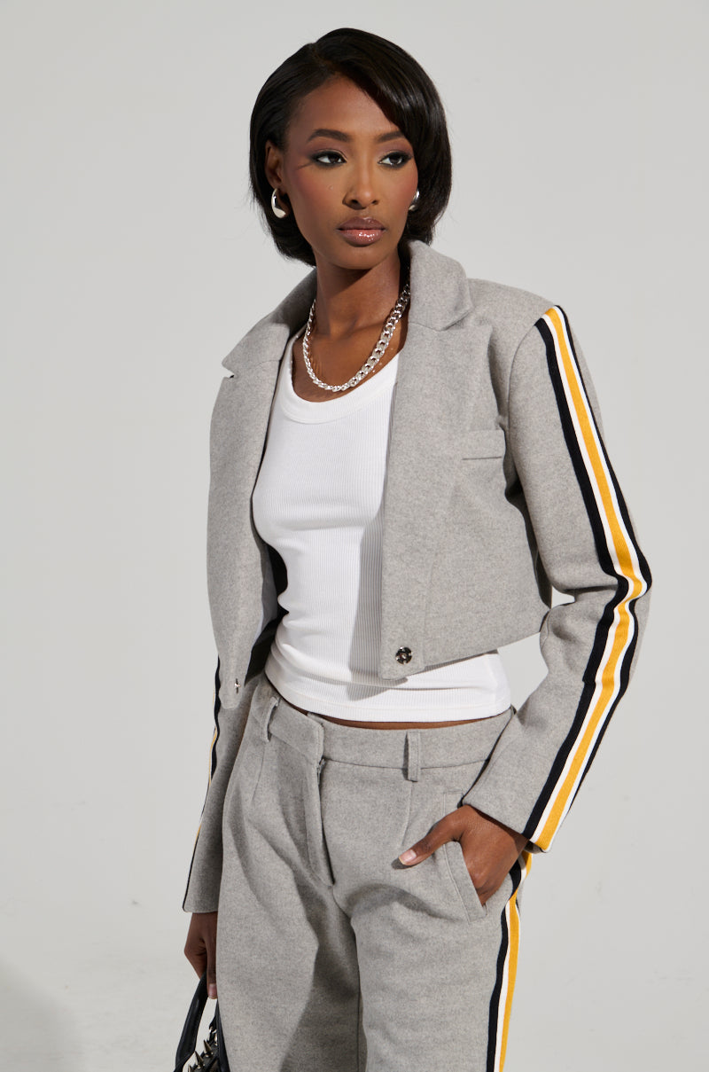 A woman exudes business-casual elegance in an AKIRA Label STAY WITH ME CROPPED BLAZER, featuring gray fabric with yellow and black striped sleeves and sides. She pairs the blazer with a white top, hoop earrings, and a silver necklace, standing confidently with one hand in her pocket.