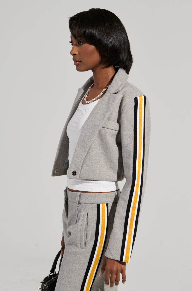 A person wearing the STAY WITH ME CROPPED BLAZER, featuring black, yellow, and white stripes on the sleeves, stands sideways. They embody a business-casual style with a white shirt underneath and hold a black handbag. A necklace adorns their neck, while their shoulder-length straight hair adds elegance against the plain background.