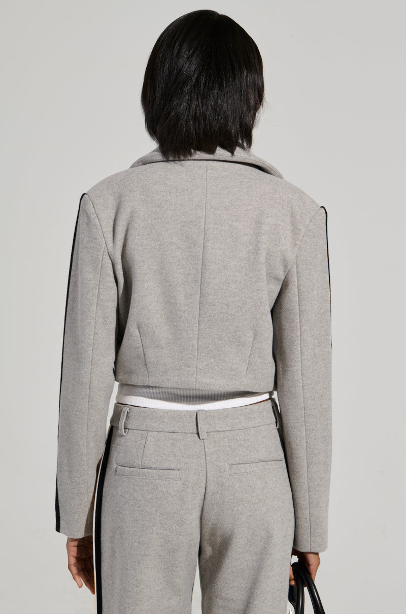 A person with short, dark hair faces away from the camera, wearing an AKIRA Label business-casual ensemble. The light gray STAY WITH ME CROPPED BLAZER and matching pants feature black stripes. In their left hand, they hold a small black bag.