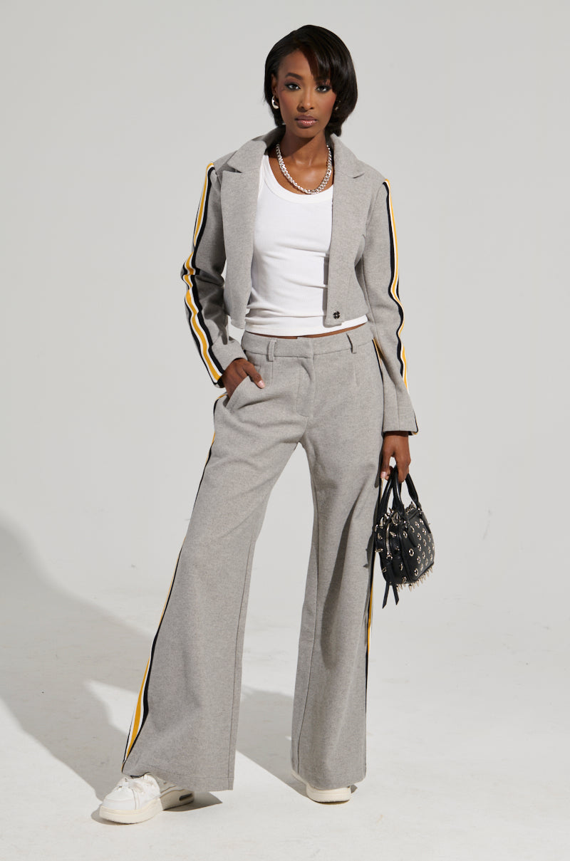 A woman radiates business-casual elegance in an AKIRA Label STAY WITH ME CROPPED BLAZER gray pantsuit featuring black and yellow striped sleeves. Her ensemble is completed with a white T-shirt, white sneakers, and a sleek black purse. She showcases her short hair and a stylish necklace against the light-colored background.
