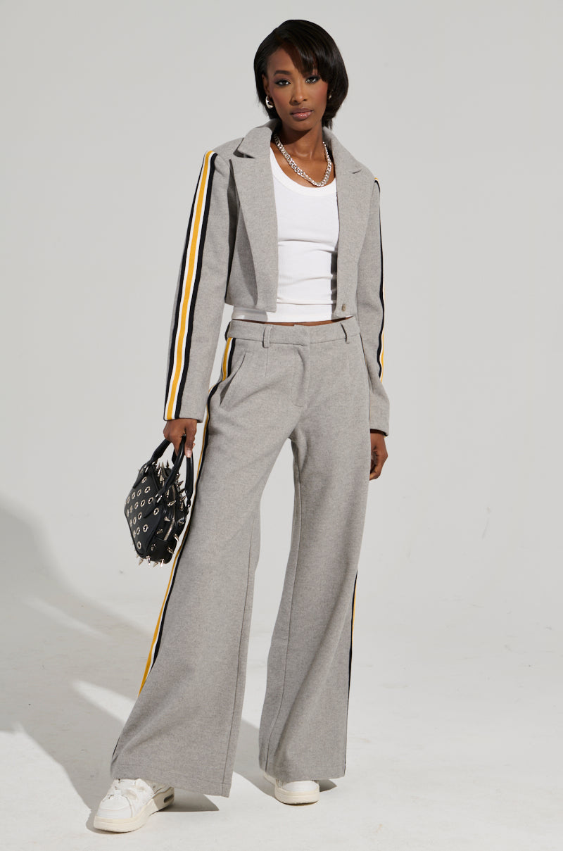 Dressed in a chic STAY WITH ME CROPPED BLAZER from the AKIRA Label collection, featuring gray fabric with eye-catching yellow and black striped details on the sleeves and pants, a person exudes confidence. This business-casual ensemble includes a white top, paired with clean white sneakers and complemented by a stylish black bag adorned with white patterns.