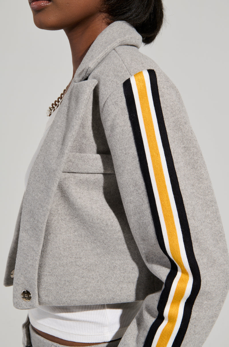 A person wearing the STAY WITH ME CROPPED BLAZER by AKIRA Label, featuring gray fabric with black, white, and yellow striped detailing on the sleeve, seamlessly incorporates it into a business-casual outfit with a white top. Their short hairstyle highlights a visible earring against the plain background.