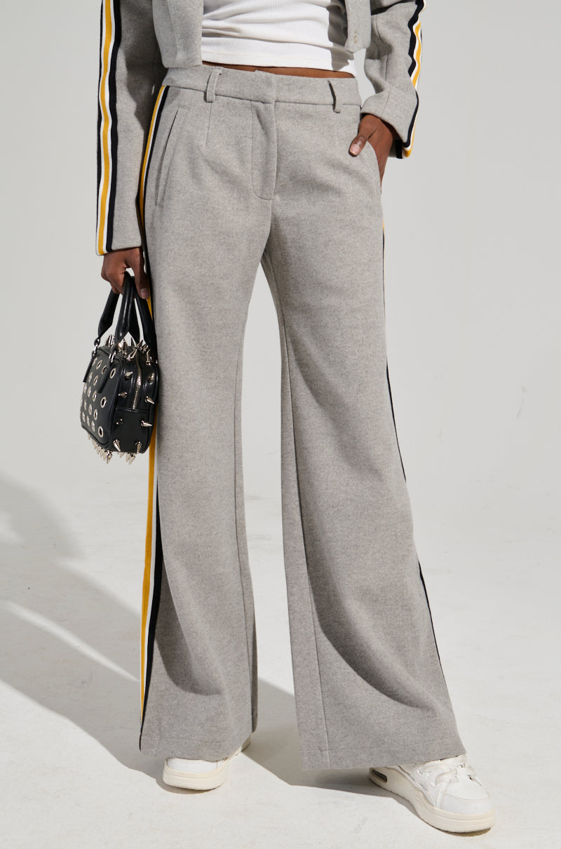 A person dressed in the STAY WITH ME WIDE LEG PANT, featuring gray fabric with black and yellow color block stripes on the sides. This outfit is paired with a coordinating jacket and white sneakers. They are carrying a studded black bag.