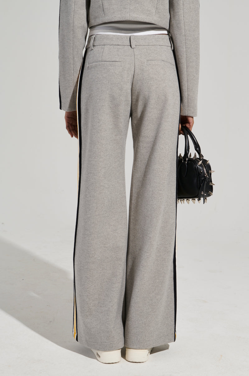 A person wearing the STAY WITH ME WIDE LEG PANT in gray, featuring black and yellow color block stripes, stands with their back to the camera. They carry a small black handbag adorned with metal studs. The background is plain and neutral.