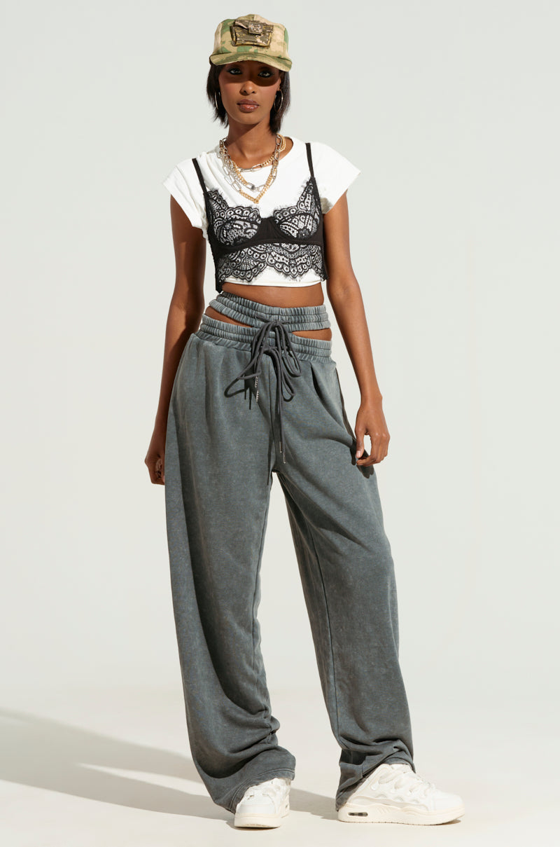 A person stands confidently wearing a camo cap, sunglasses, and a white T-shirt with a black lace bralette over it. They sport the NEXT UP IS ME SWEATPANT in gray, featuring a wide-leg silhouette and dual shirred elastic waistband. Layered necklaces accent the look, and their hands rest in their pockets.