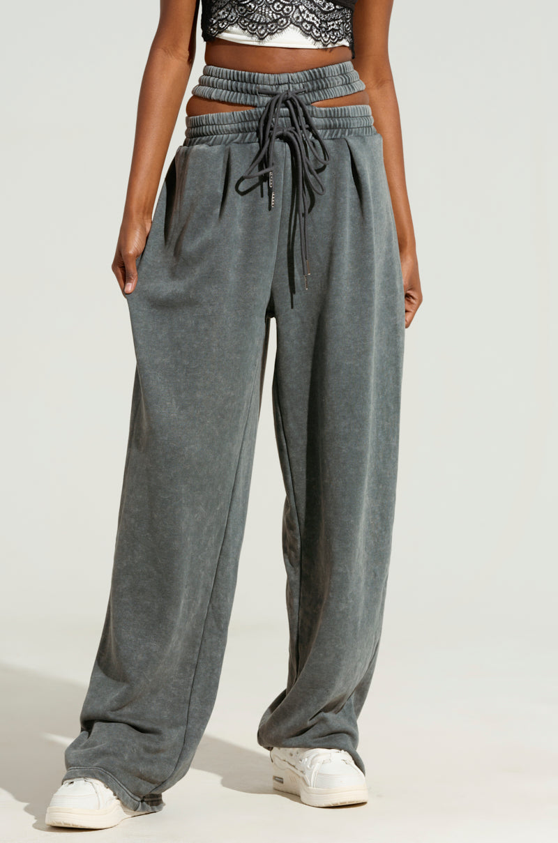 A person wearing the "NEXT UP IS ME" high-waisted, wide-leg charcoal gray sweatpants featuring a dual shirred elastic waistband, paired with a black lace crop top. They are standing against a neutral background and wearing white sneakers.