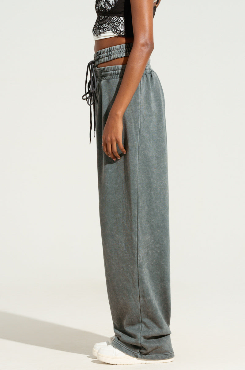 A person wearing the NEXT UP IS ME SWEATPANT in dark gray, featuring a high-rise design and wide legs with an elastic waistband and drawstring, stands in profile against a plain background. They pair the sweatpants with a black patterned crop top and white sneakers.