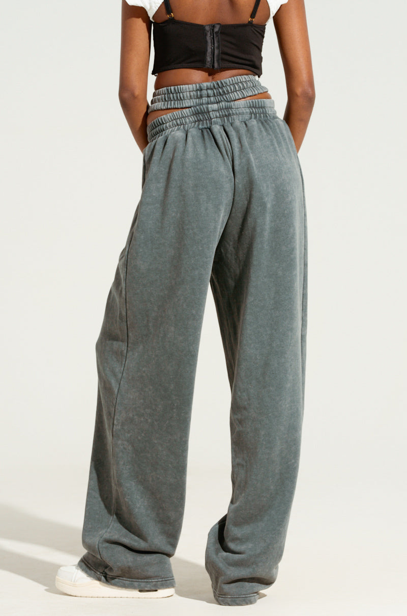 The individual is dressed in the NEXT UP IS ME SWEATPANT, featuring a dual shirred elastic waistband in a high-rise gray design, paired with a black strappy top and white sneakers. They are standing against a plain background with their hands on their hips.