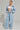 A person stands confidently in a fashion-forward outfit, flaunting the HONEY HONEY WIDE LEG PANTS in a high-rise light blue velour design, accompanied by a matching jacket and white crop top. They accessorize with hoop earrings and layered necklaces, showcasing a tattoo on their midriff.