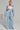 A person stands confidently wearing the HONEY HONEY WIDE LEG PANTS, a light blue cotton blend knit outfit featuring high-rise, wide-leg pants paired with a cropped hoodie. They also sport a white bandeau top underneath, multiple necklaces, and have their hair styled in a sleek updo.