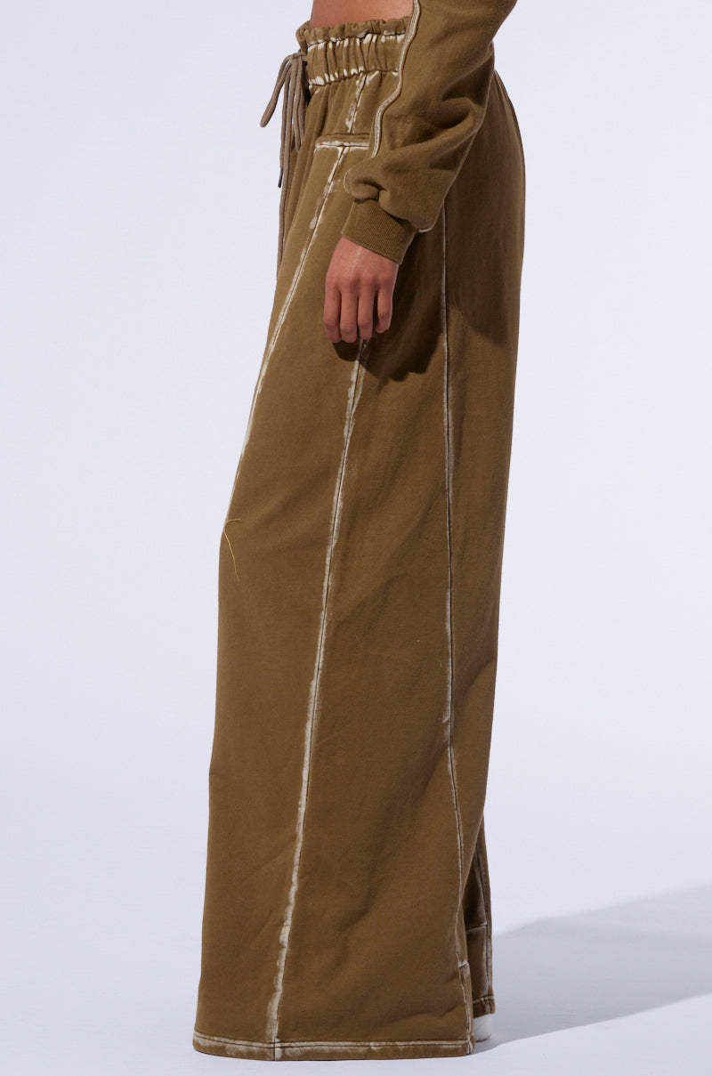 A person is dressed in a pair of the STREET TALK WIDE LEG PANT in khaki, featuring a high rise shirred waistband, visible white stitching, and a drawstring waist. The pants are complemented by a matching long-sleeve top. The backdrop is light and plain, showcasing the individual's side profile from mid-torso to feet.