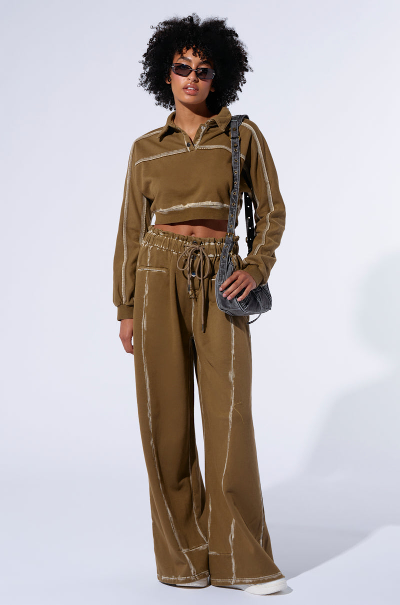 A person with curly hair, wearing sunglasses, poses confidently in a brown long-sleeve crop top and matching STREET TALK WIDE LEG PANT with a high rise shirred waistband. They have a shoulder bag slung across their body. The background is plain white.