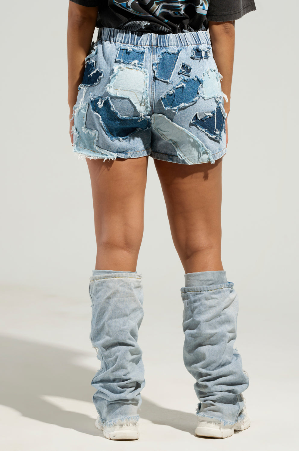 A person is standing with their back to the camera, wearing BAD IDEA DENIM PATCHWORK SHORTS characterized by a high-rise design and elastic waistband. The shorts showcase various patches and frayed edges, paired with denim leg warmers. They have styled it with a dark top and white shoes against a plain background.