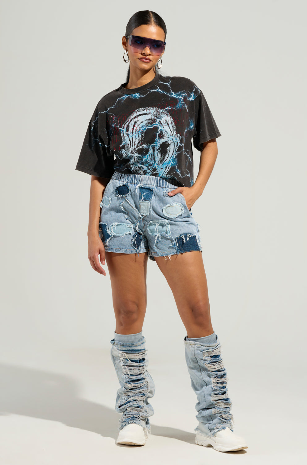 A person confidently stands against a neutral background, showcasing a black graphic T-shirt, the BAD IDEA DENIM PATCHWORK SHORTS with an elastic waistband, denim leg warmers, white shoes, and stylish sunglasses.