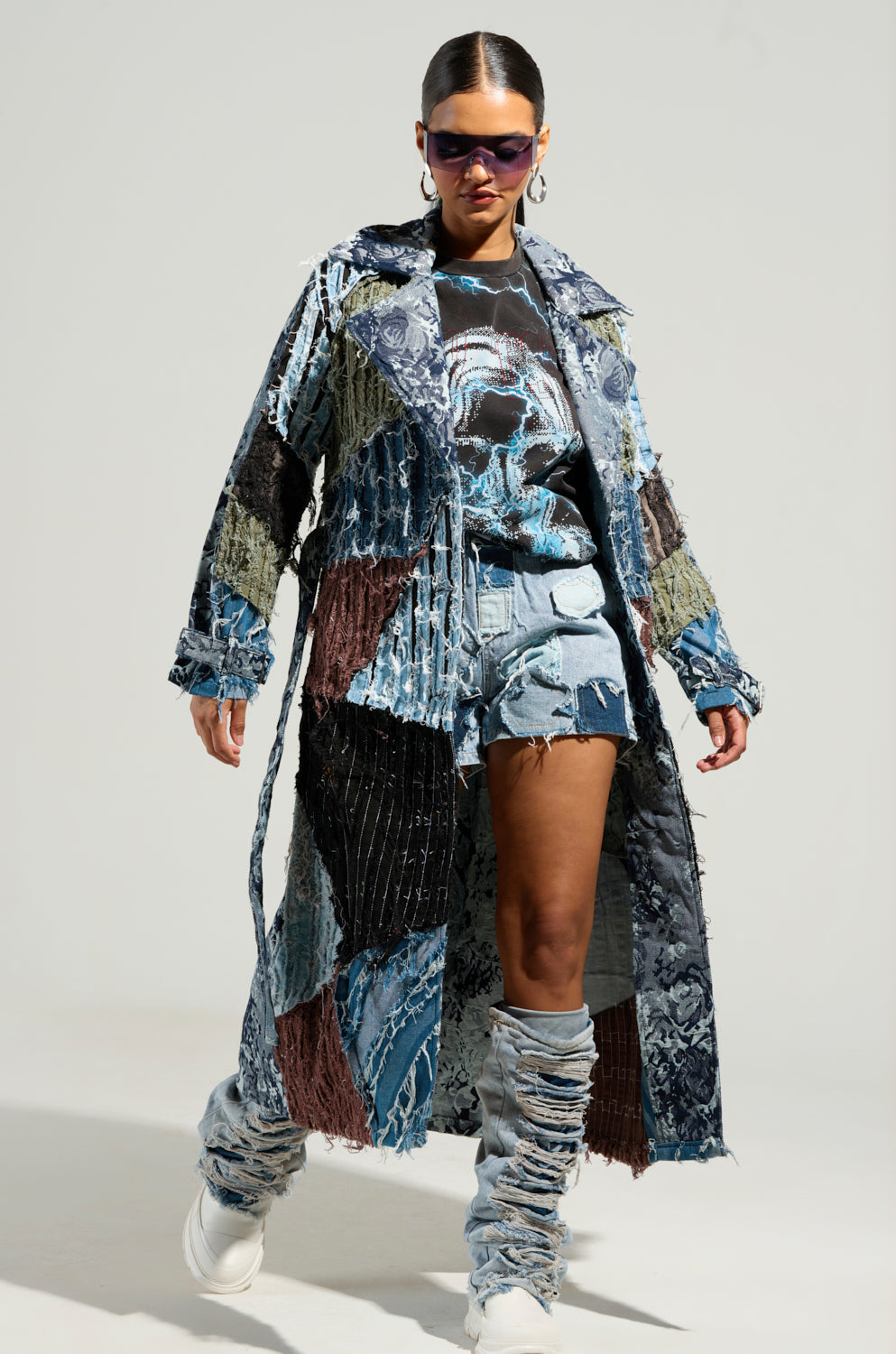 Sporting a striking patchwork denim ensemble, the individual models the BAD IDEA DENIM PATCHWORK SHORTS, complete with an elastic waistband. The outfit is complemented by a long coat and leg warmers showcasing a vivid abstract pattern. They finish the look with sunglasses and white shoes, exuding confidence against a plain backdrop.