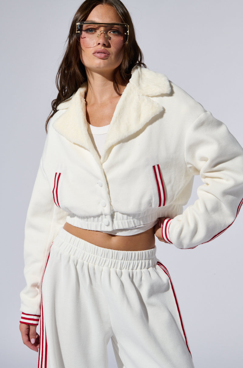 A woman poses confidently, dressed in matching white fleece sweatpants and a Lula Waffle Knit Bomber adorned with varsity-inspired stripes on the cuffs and sides. She wears a white top underneath and stylish tinted sunglasses, her hands resting on her hips against a neutral background.