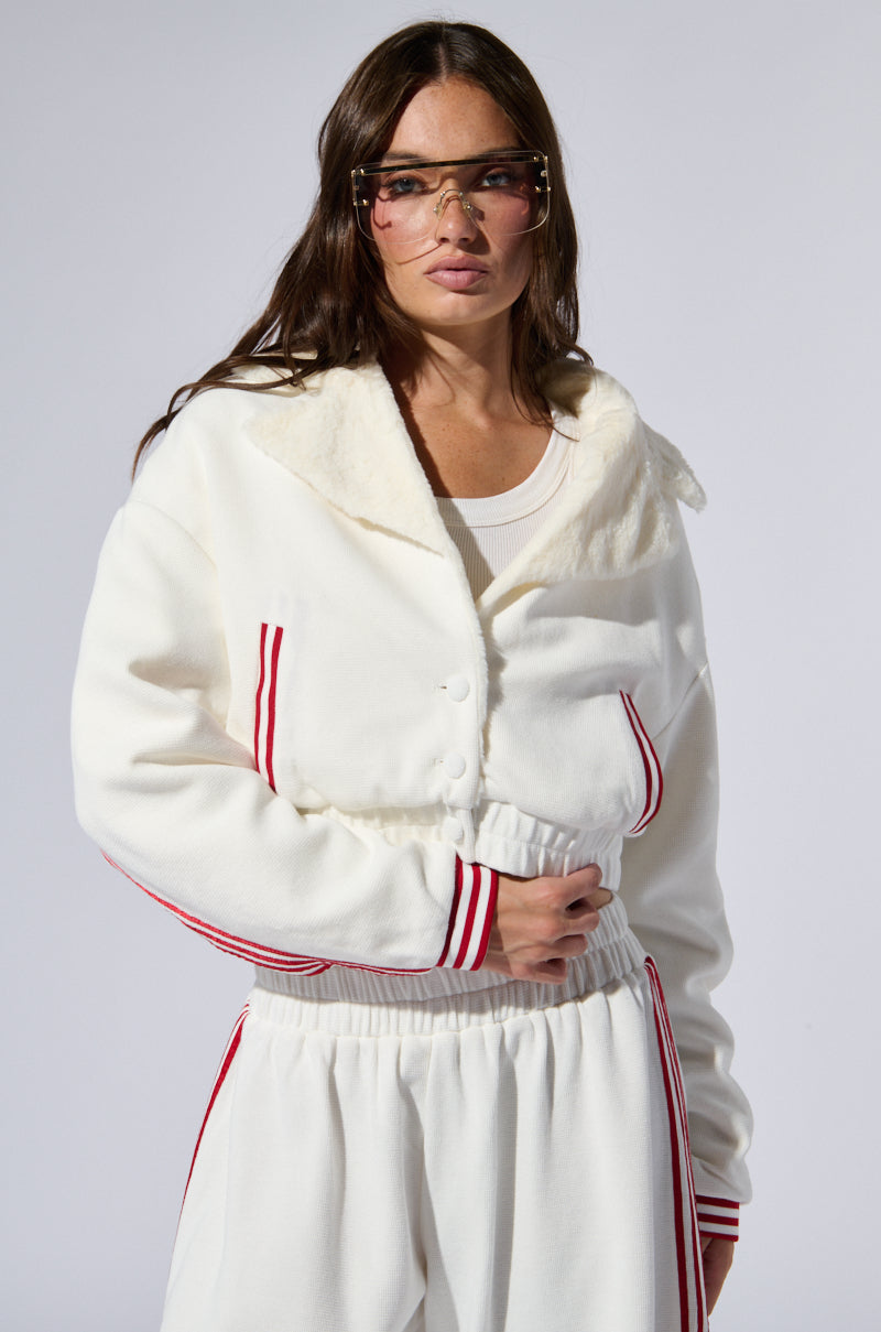 A woman with long brown hair wears transparent visor-style glasses, the LULA WAFFLE KNIT BOMBER jacket featuring varsity-inspired stripes and a faux fur collar, and matching white pants. She stands against a plain background, with one hand on her hip.