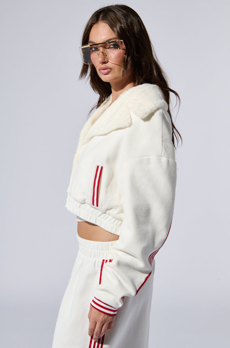 A person stands in a side profile pose wearing the Lula Waffle Knit Bomber, a white cropped hoodie featuring varsity-inspired stripes on the sleeves, along with matching white sweatpants adorned with red stripes. They have long brown hair and are wearing large, square, clear glasses. The background is plain and light-colored.