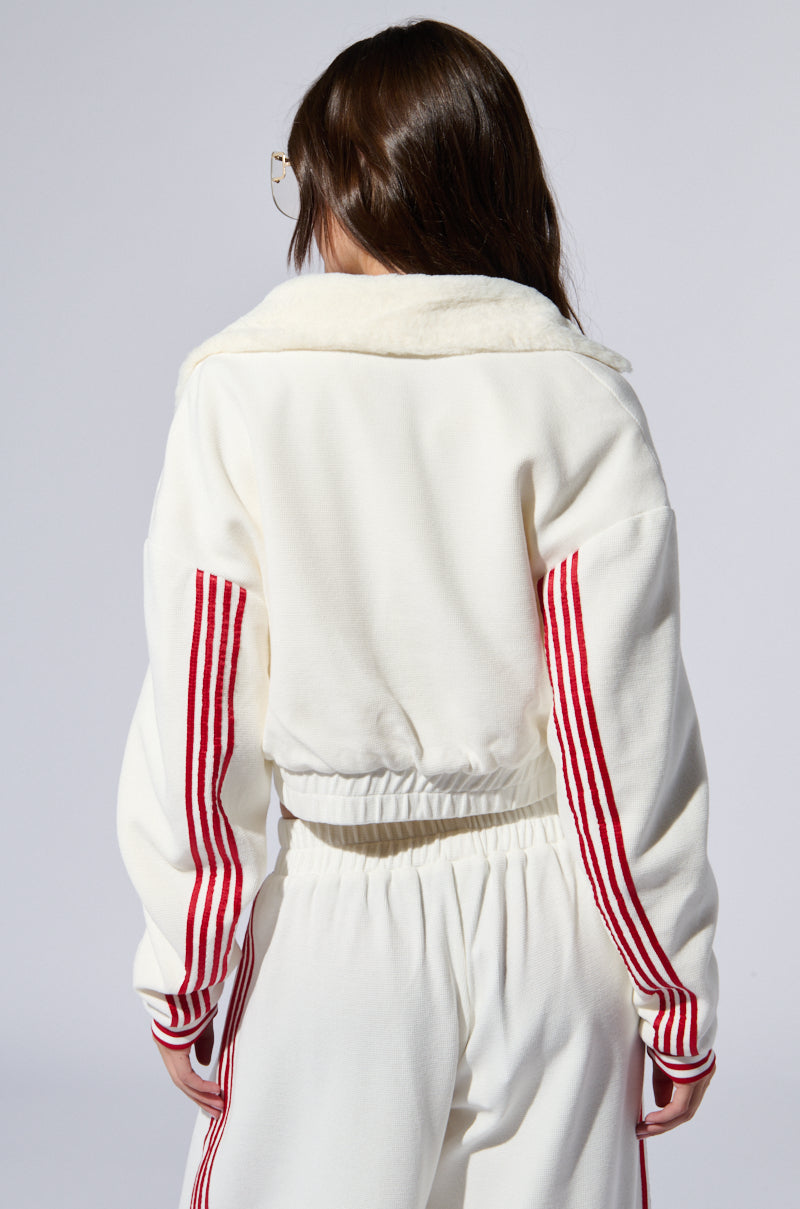 A person with long brown hair, wearing glasses, is shown from behind. They are dressed in the LULA WAFFLE KNIT BOMBER, a matching white track suit featuring varsity-inspired stripes on the sleeves and pants. The bomber has a faux fur lining collar, and the background is plain gray.