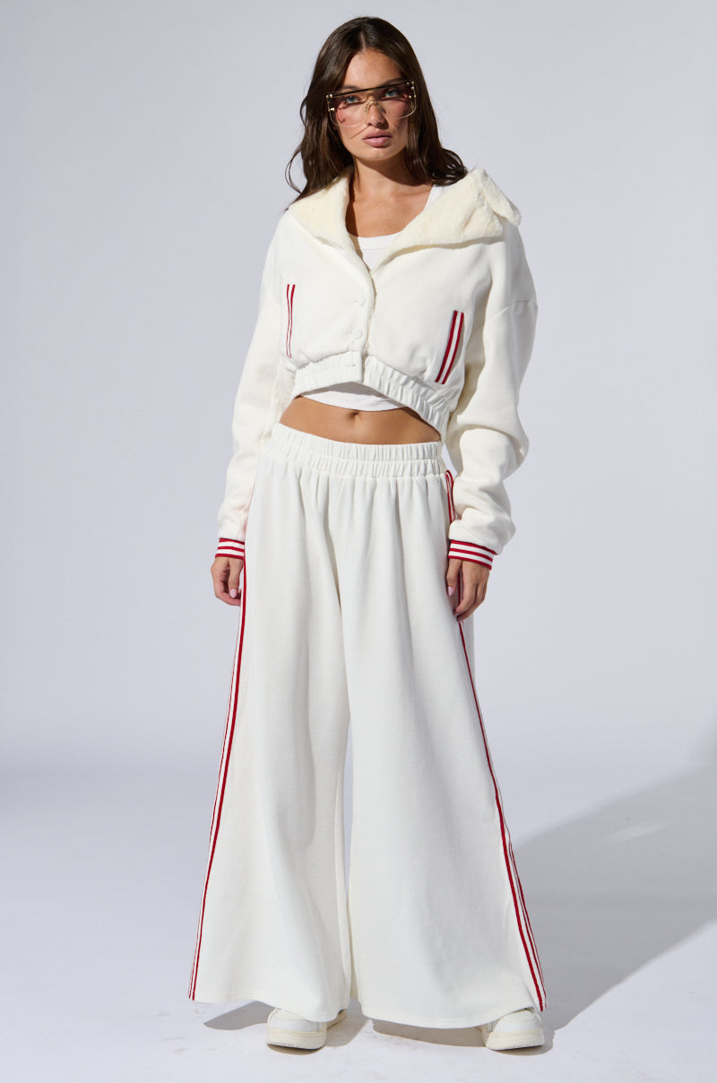 A woman stands against a plain backdrop wearing the LULA WAFFLE KNIT BOMBER, a coordinated white outfit featuring varsity-inspired red stripes. The ensemble includes a cropped, hooded jacket with faux fur lining and wide-legged pants. She has long hair and is wearing glasses.