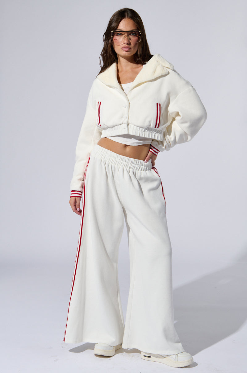 A person with long hair stands confidently, wearing the stylish white LULA WAFFLE KNIT BOMBER paired with wide-legged pants featuring varsity-inspired stripes. They are also sporting sunglasses, giving a chic and modern vibe against a plain white background.