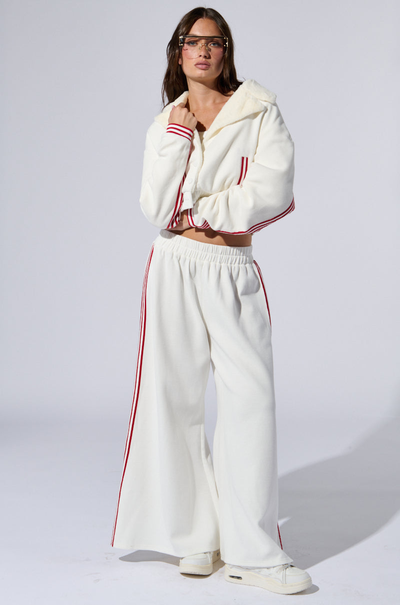 A person with long hair stands confidently against a plain background, wearing a white LULA WAFFLE KNIT BOMBER with varsity-inspired red stripes along the sleeves and pants. They accessorize with glasses and white sneakers, showcasing a pensive expression and relaxed pose.