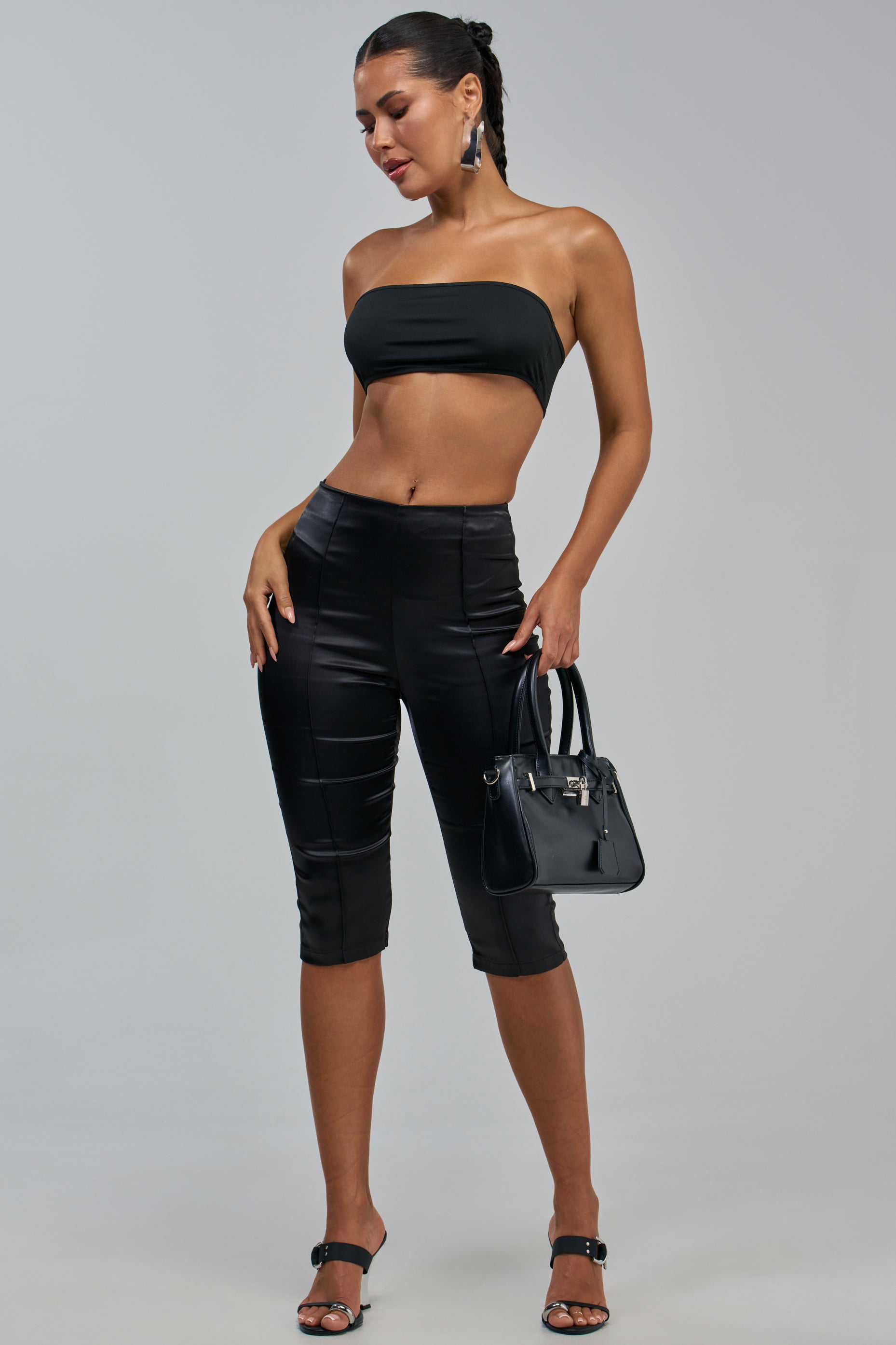 A woman stands against a plain gray background wearing the HIT THE LIGHTS SATIN CAPRI in black, paired with a black bandeau top, black heeled sandals, and a black handbag. Her hair is pulled back.