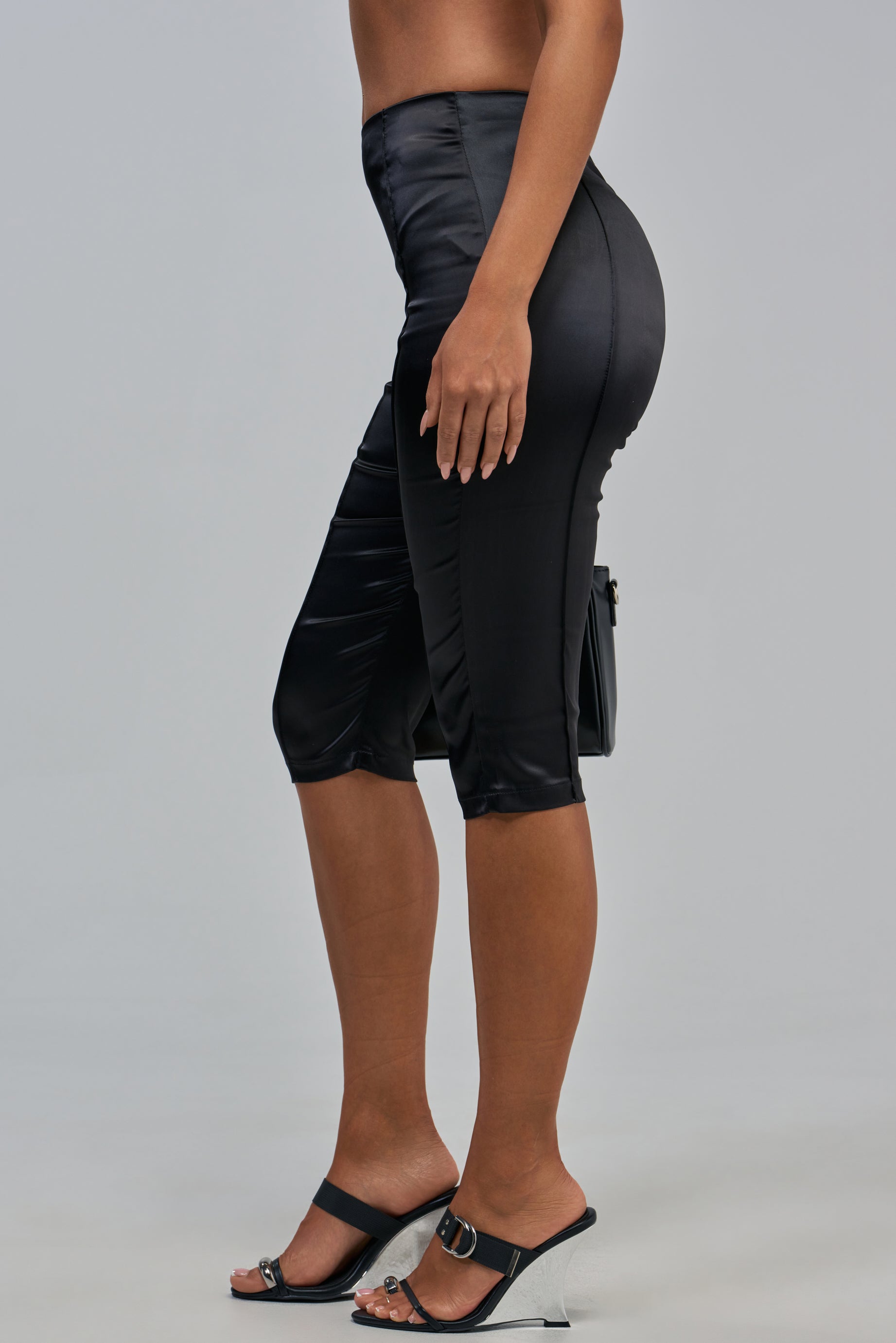 A person wearing the HIT THE LIGHTS SATIN CAPRI with a high-rise fit, paired with black high-heeled sandals and carrying a small black handbag, shown from the side against a plain background.
