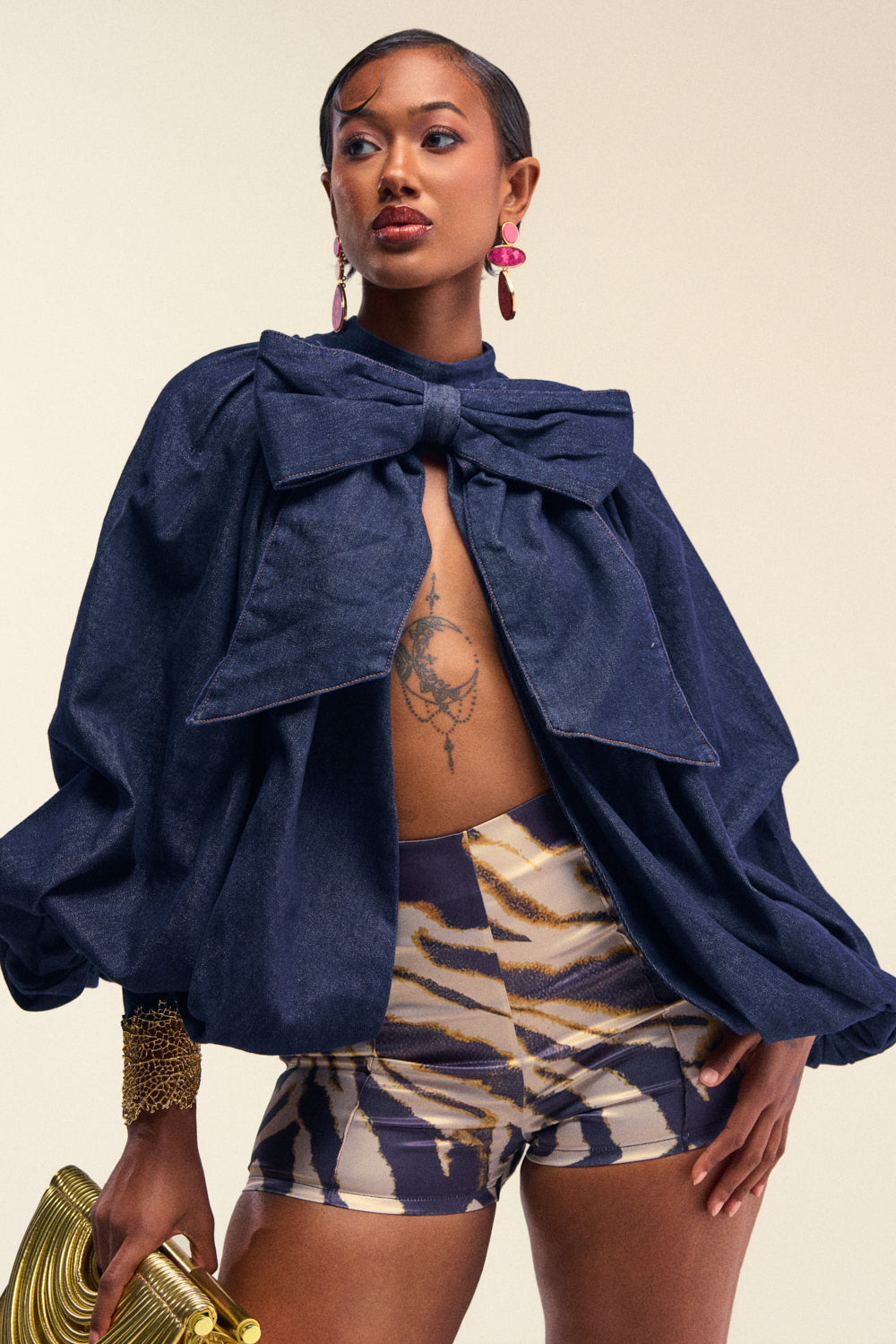 A confident woman poses in an oversized dark denim jacket with a large bow, THE BEST TIGER SATIN SHORT high-rise shorts, a gold bracelet, and red earrings, holding a gold clutch. She has a chest tattoo and slicked-back hair—perfect for bold festival styling.