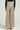 A person stands facing away, highlighting the FEELING GODLY PLEATED HIGH WAIST PANT IN GOLD paired with a black top. Their open-toed heels accentuate the elegant flowy wide-leg silhouette, set against a simple off-white backdrop that enhances their striking ensemble.