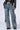 A person wearing the EVERY LITTLE THING SHE DOES DENIM PANT—featuring a loose fit, high-rise waist, multiple cargo pockets, and straps—along with a black leather jacket and black chunky shoes. The person's face is not visible. The background is plain light gray.