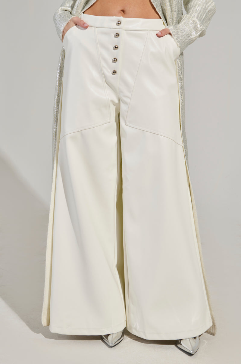 A person wearing the ASHLYN FAUX LEATHER PANTS in white, which are mid-rise with a wide-leg cut and feature buttons down the front. These stylish pants, complete with functional pockets, complement a long shimmering coat. The individual stands with hands comfortably nestled in the pockets while silver pointed shoes peep out from beneath the pant legs, enhancing a sleek silhouette. 2