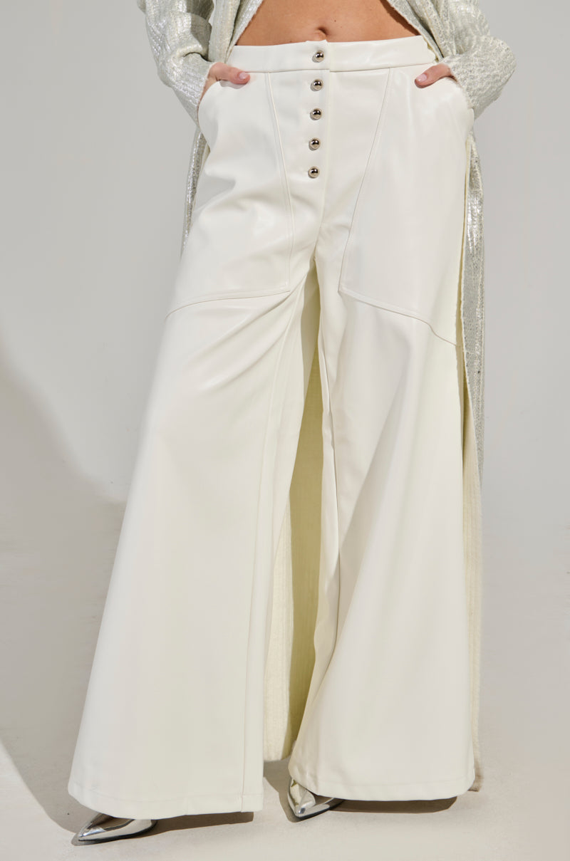 A person is dressed in the ASHLYN FAUX LEATHER PANT IN WHITE, featuring wide legs and a high waist accentuated by large buttons. The ensemble includes a shimmering silver coat, with silver pointed shoes visible beneath the wide-legged silhouette, all set against a gray backdrop with the person cropped above the waist. 0